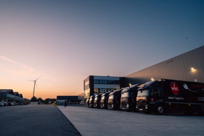 Des camions électriques alignés devant le centre de distribution, avec une éolienne en arrière-plan.